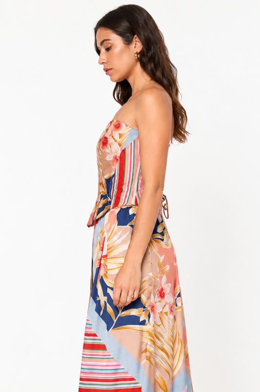 A woman modeling a strapless maxi dress with a multicolor abstract print featuring stripes, floral motifs, and leaf patterns in pink, blue, red, beige, and orange, shown from a side profile against a plain white background.