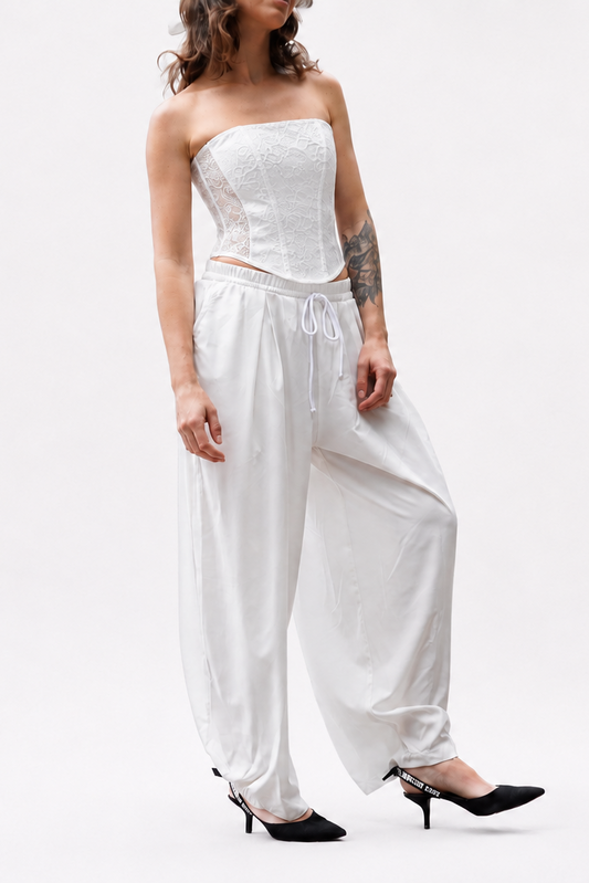 A model wearing a white strapless corset-style top and matching loose white harem-style pants, paired with black pointed heels, against a plain white studio background.