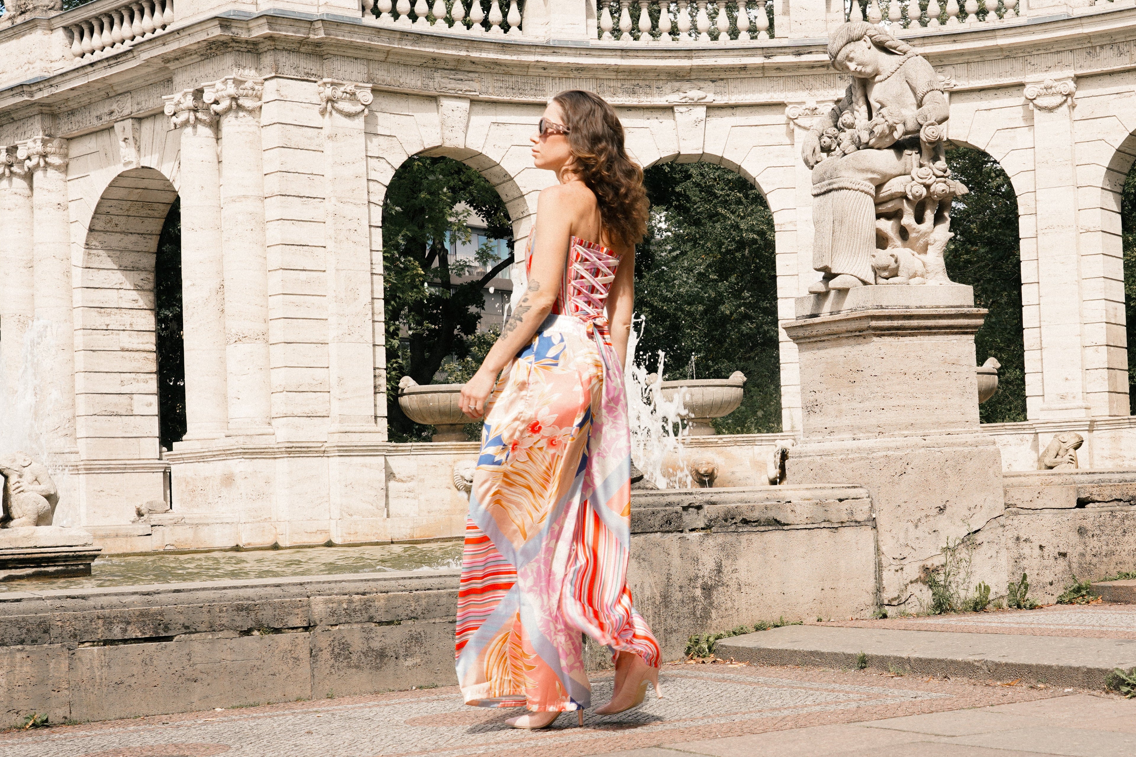 A woman wearing a colorful floral print maxi dress walks past a classical stone fountain with arches and statues in a park setting.
