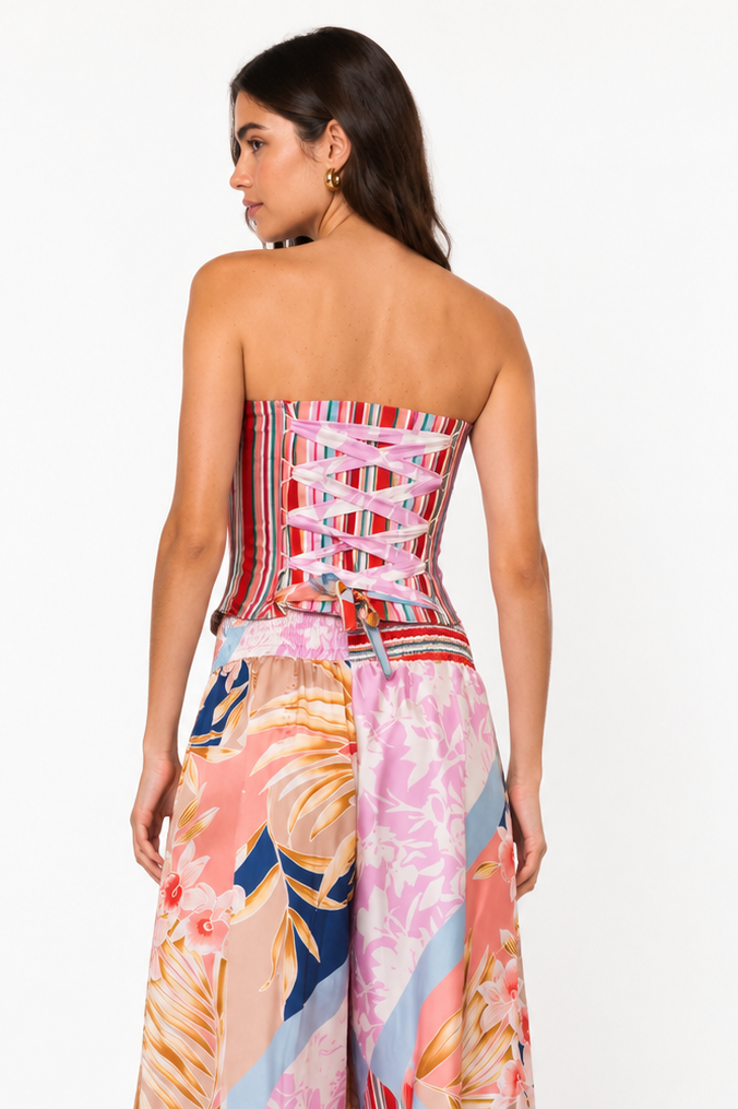 A woman modeling a strapless maxi dress from the back, featuring a corset-style waist with lace-up detailing and wide, flowing skirt panels in a pink, orange, blue, and red tropical and striped print.