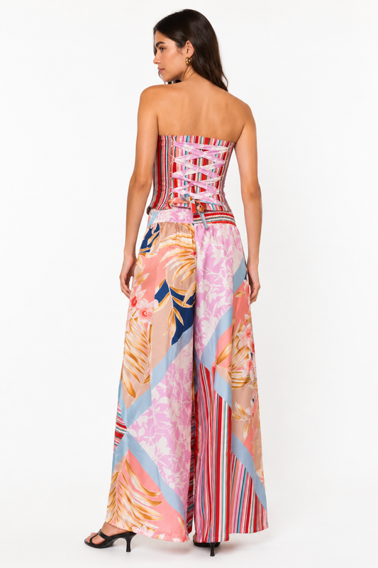 A woman modeling a strapless maxi dress from the back, featuring a corset-style waist with lace-up detailing and wide, flowing skirt panels in a pink, orange, blue, and red tropical and striped print.