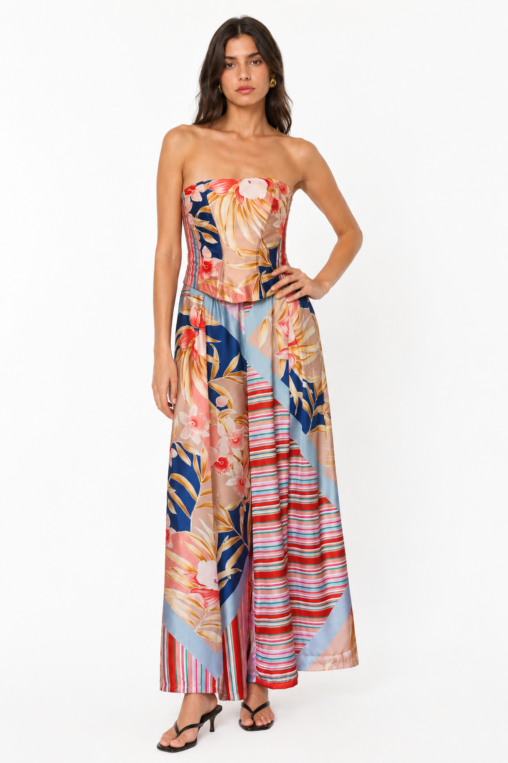 A woman models a strapless maxi dress with a vibrant tropical floral print and striped panels in shades of blue, red, pink, and beige against a plain white studio background.