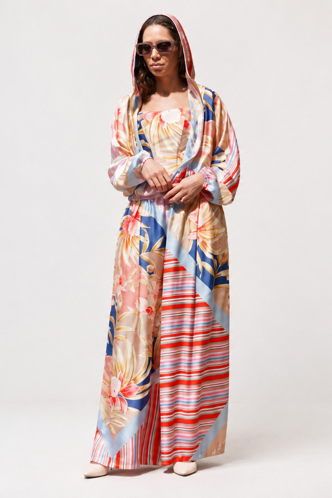 A woman modeling a long, loose, kaftan-style dress with a colorful abstract floral and striped print, paired with a matching headscarf and sunglasses, set against a plain light gray studio background.

