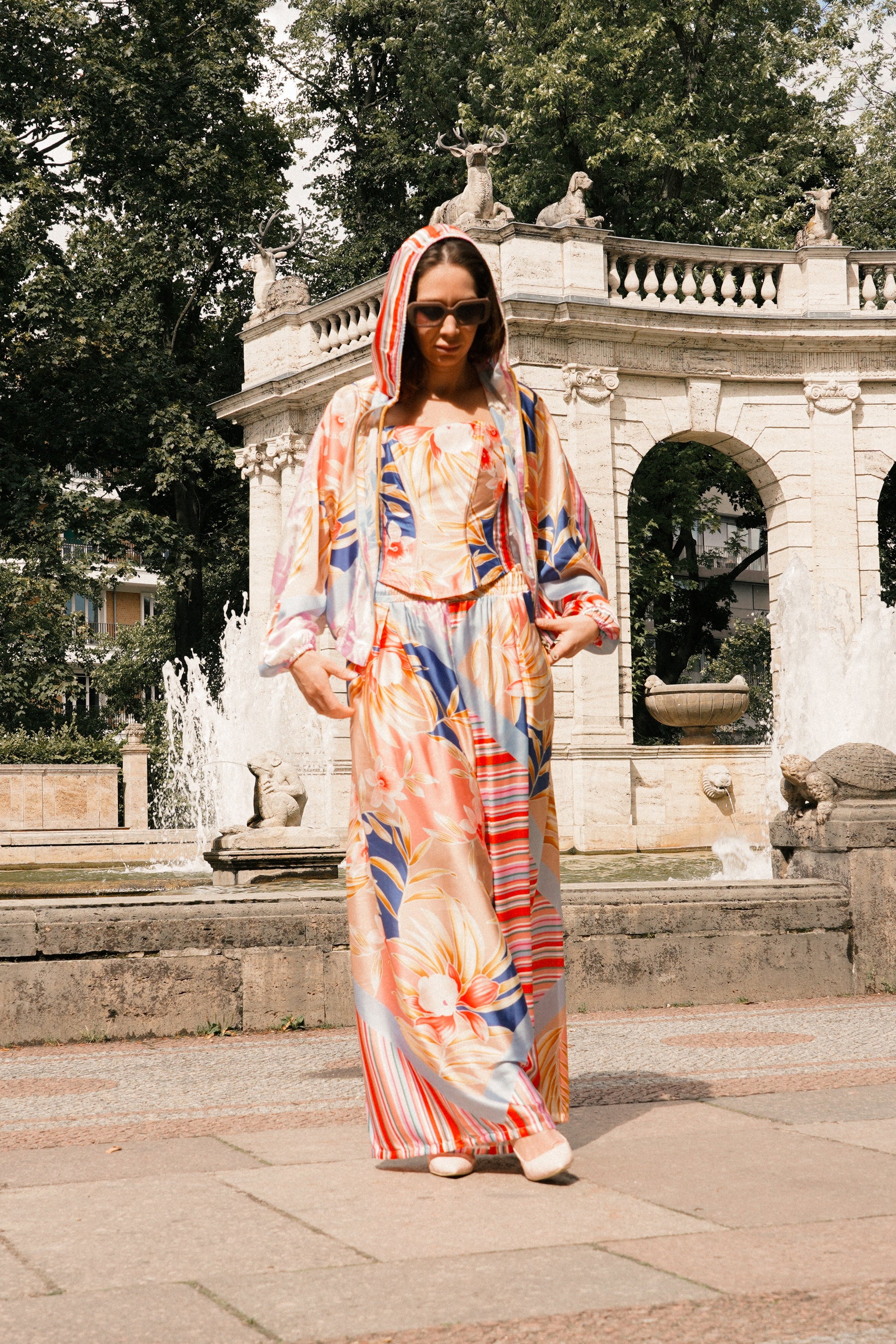 A woman wearing a colorful, patterned hooded maxi dress walks toward the camera in front of a large stone fountain and classical balustrade in a park setting.