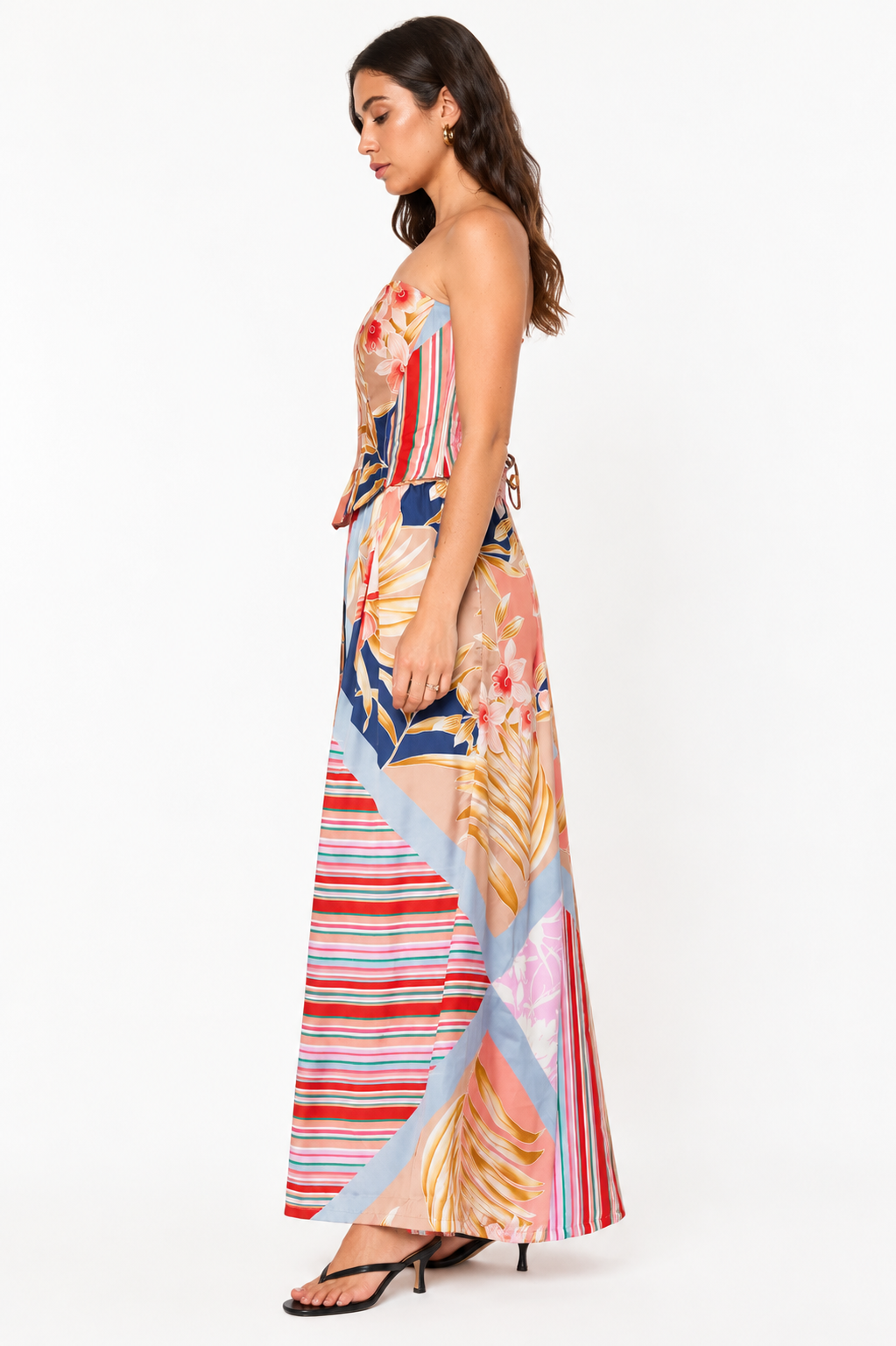 A woman modeling a strapless maxi dress with a multicolor abstract print featuring stripes, floral motifs, and leaf patterns in pink, blue, red, beige, and orange, shown from a side profile against a plain white background.