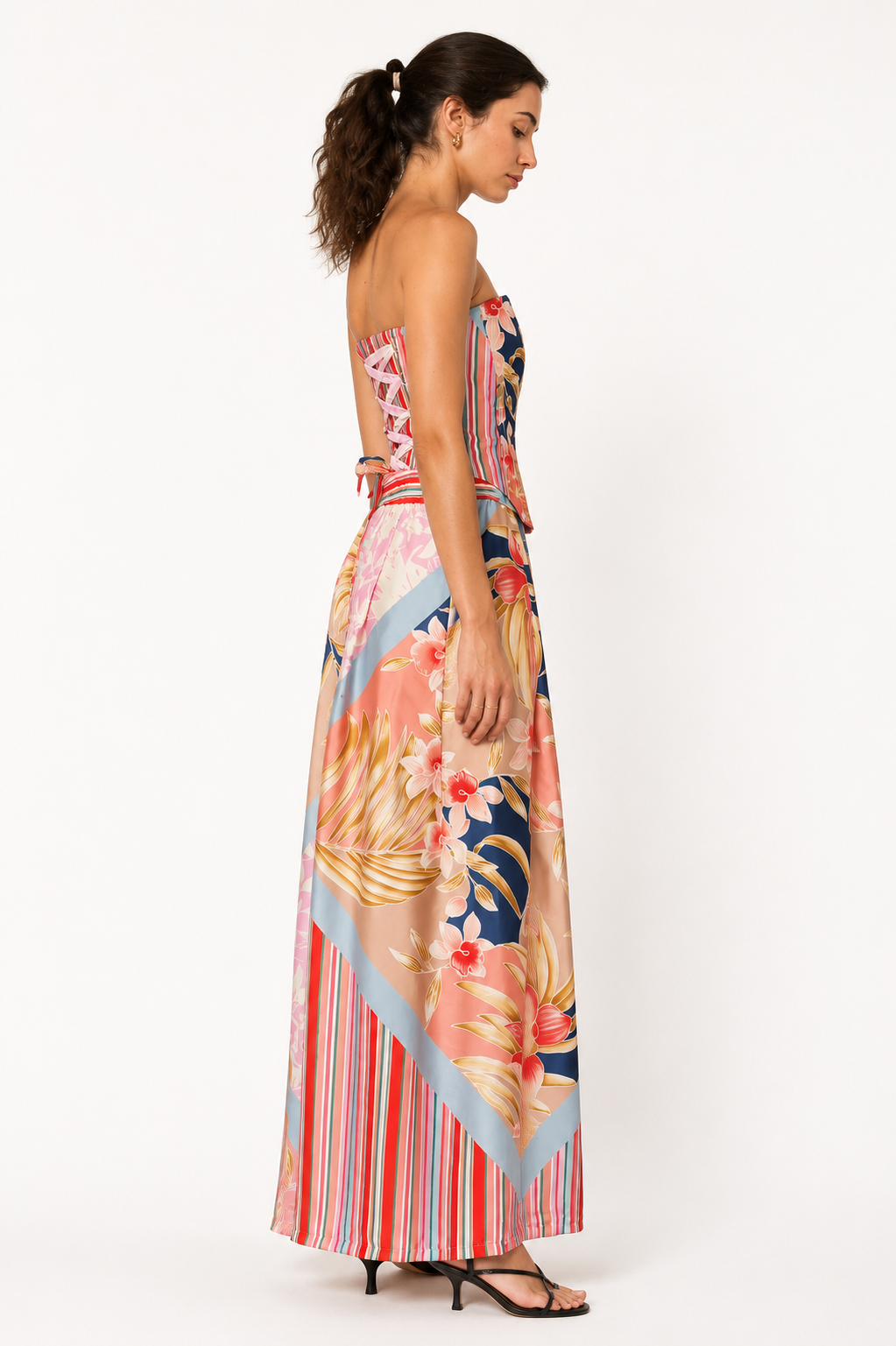 A woman modeling a strapless maxi dress with a colorful tropical floral and striped print, shown from a side-back angle against a plain white studio background.