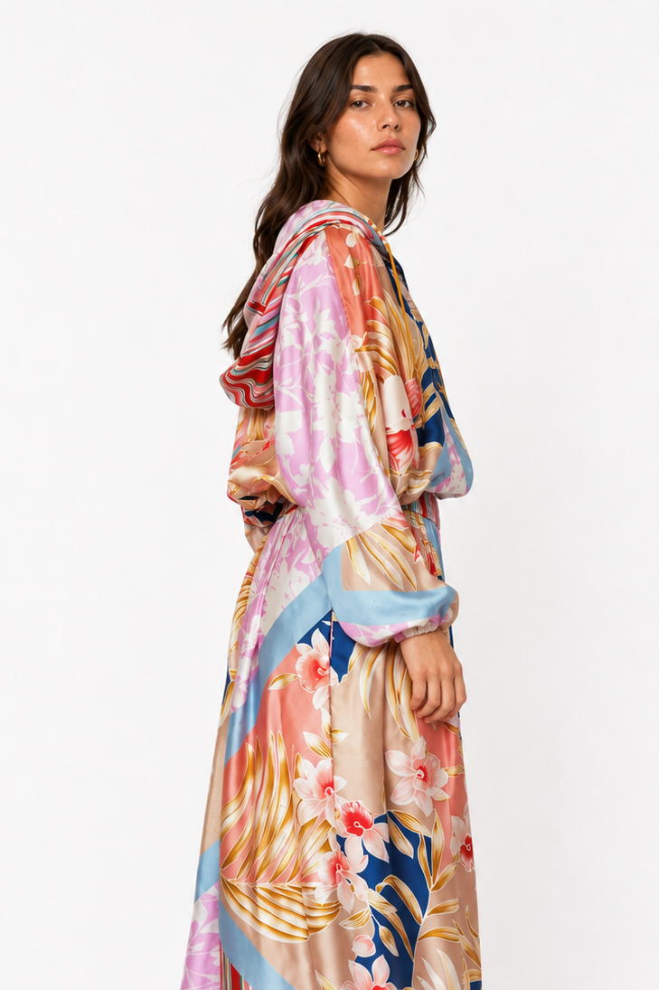 A woman modeling a long, flowing maxi dress with a vibrant multicolor abstract print featuring pink, blue, gold, and beige tones, set against a plain white studio background.