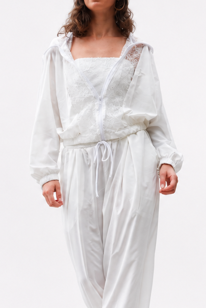 A woman modeling a white two-piece outfit consisting of a loose long-sleeve top and matching drawstring pants, paired with a white headscarf and black heels, against a solid white studio background.