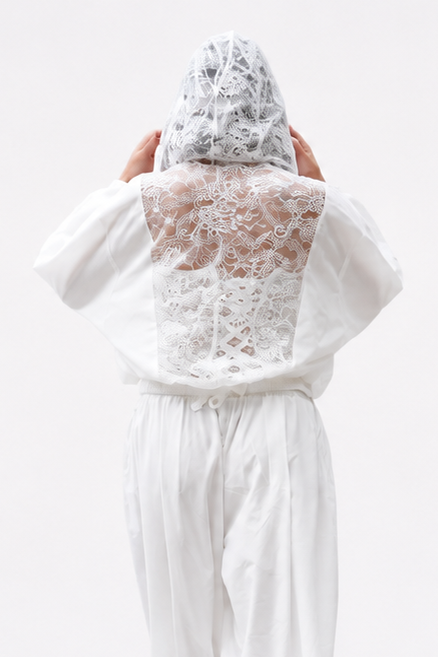 A model viewed from behind wearing a white lace-trimmed long-sleeve top and matching white wide-leg pants, with a white lace head covering and black high heels, against a seamless white studio background.