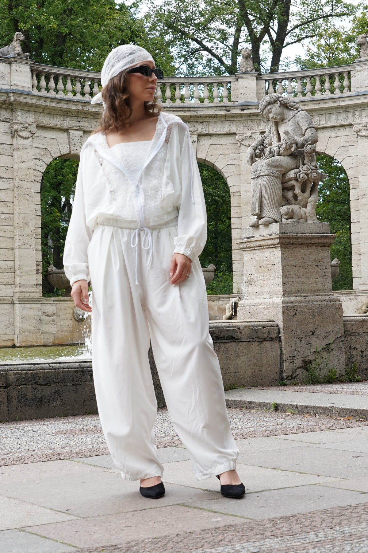 A woman wearing a white set with a matching white hat stands in front of a stone fountain and arched pavilion in a park.