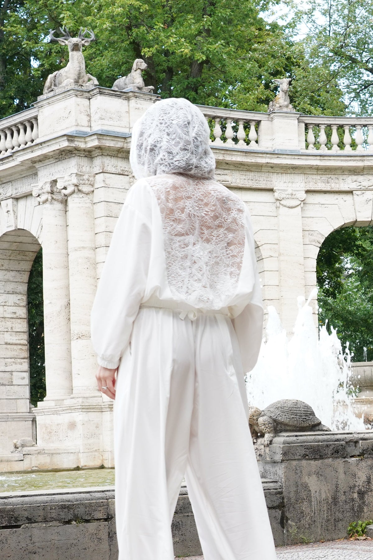 A woman viewed from behind wearing a white lace-trimmed jumpsuit and a white lace head covering, standing in front of a large classical stone arcade with arches and fountains in a park setting.