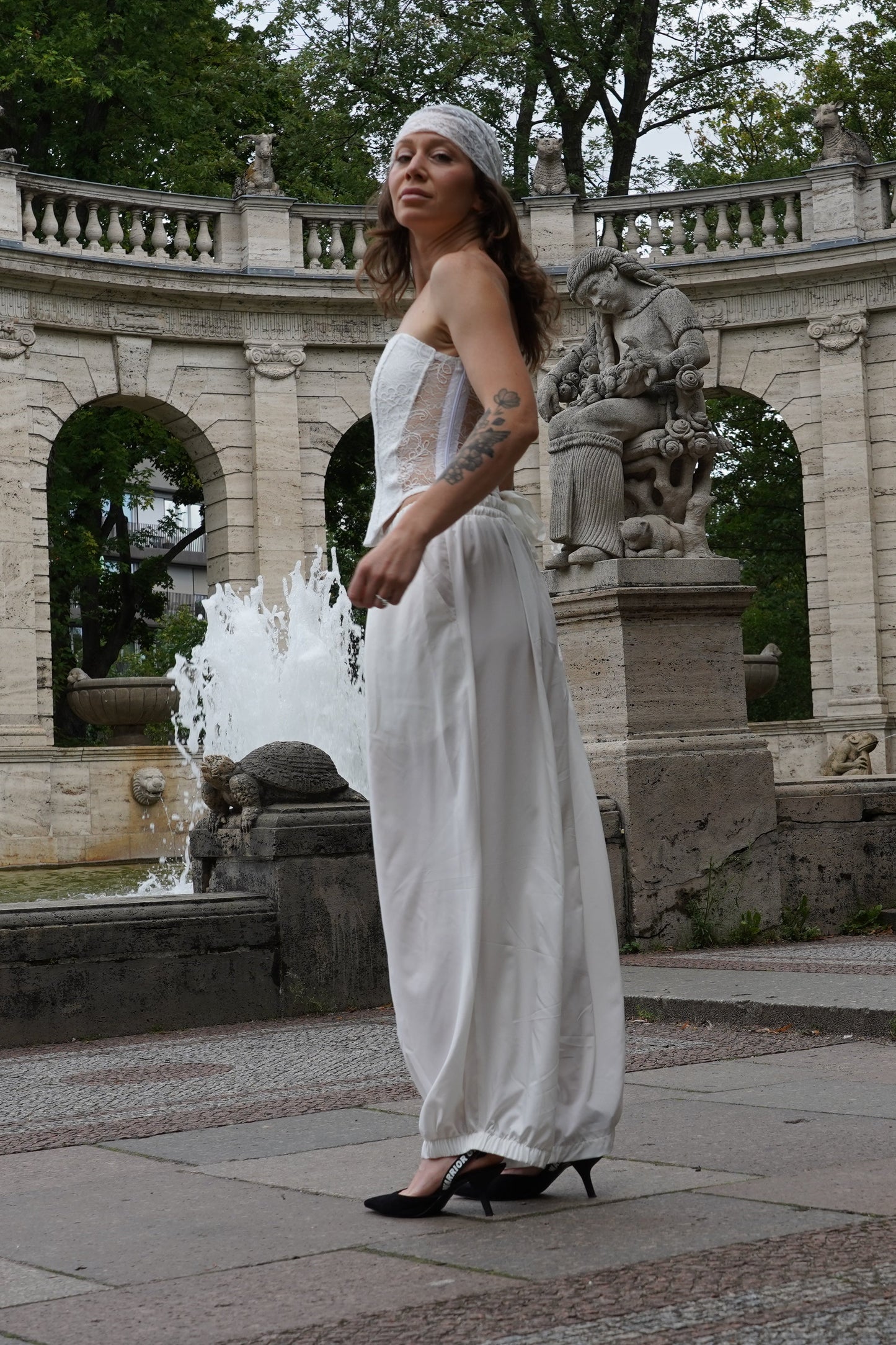 A woman wearing a white sleeveless set and a matching headband stands in front of a large stone colonnade with fountains in the background.