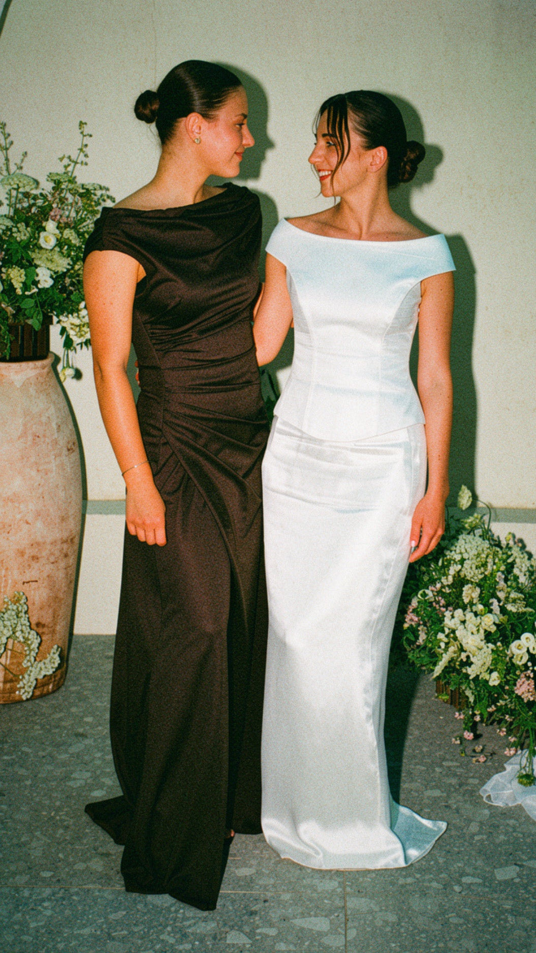 Two women standing indoors wearing formal dresses, one in a dark brown off-the-shoulder gown and the other in a white off-the-shoulder gown, with floral arrangements and draped fabric in the background.