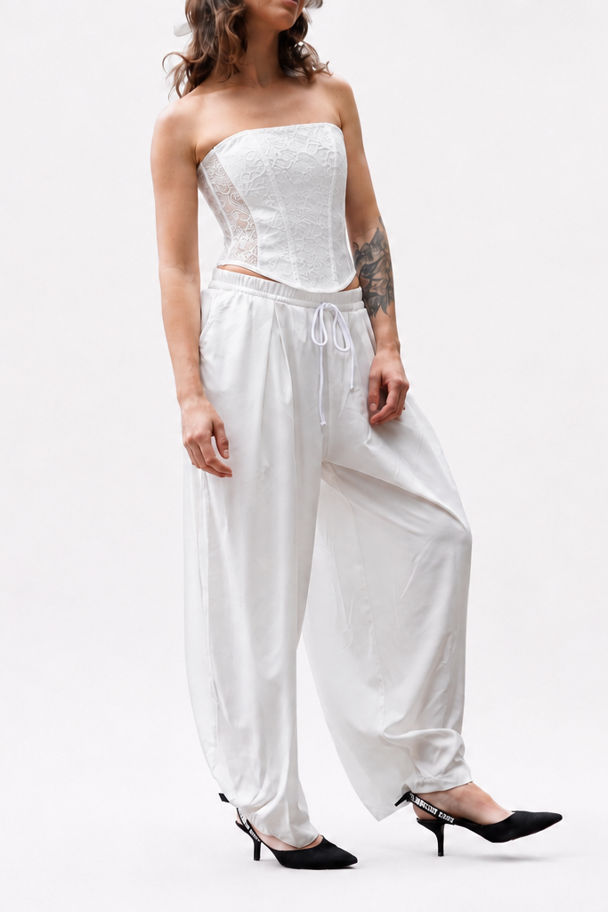 A model wearing a white strapless corset-style top and matching loose white harem-style pants, paired with black pointed heels, against a plain white studio background.